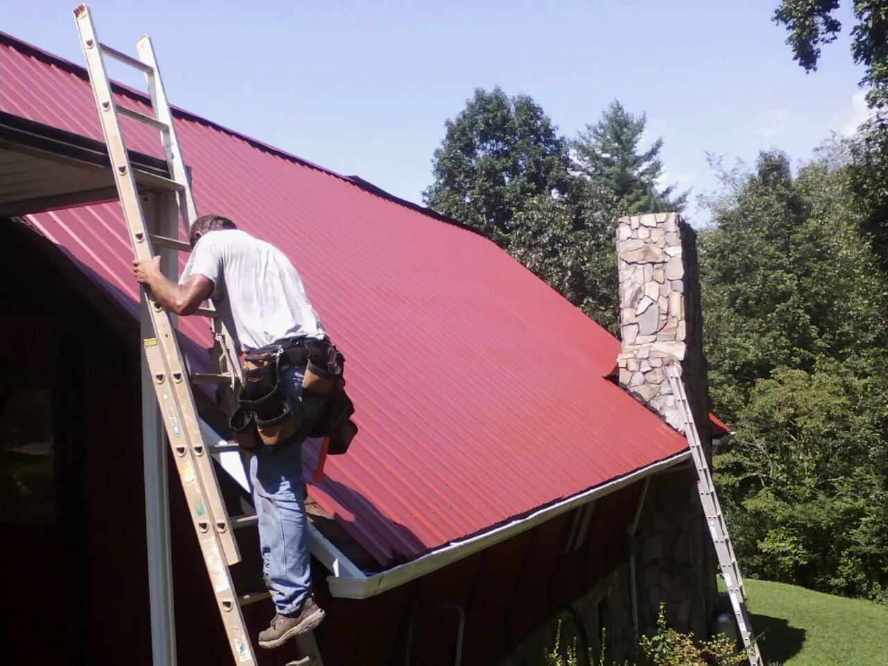 Roofer on a ladder beside a red metal roof and stone chimney — Soffit & Fascia Repair by Roofing Brothers in Gettysburg